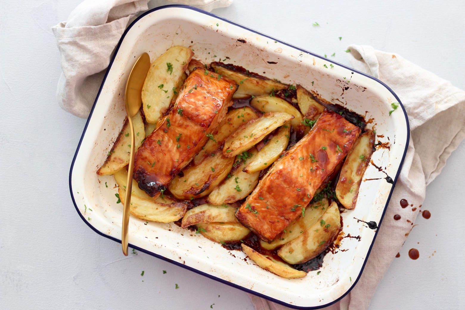 Top down image of Asian glazed salmon on a bed of crispy wedges in a baking tray with a serving spoon and tea towel