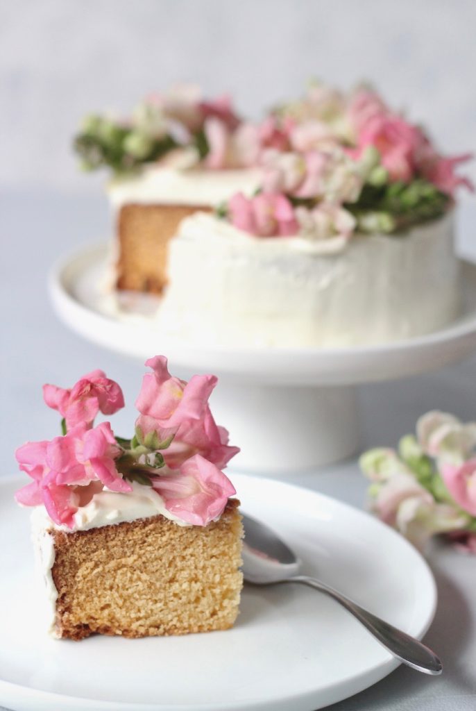 A slice of vanilla butter cake topped with pink flowers on a plate with a teaspoon and the sliced cake on a stand in the background